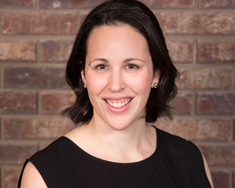 portrait of smiling woman with long brown hair wearing black shirt against brick wall