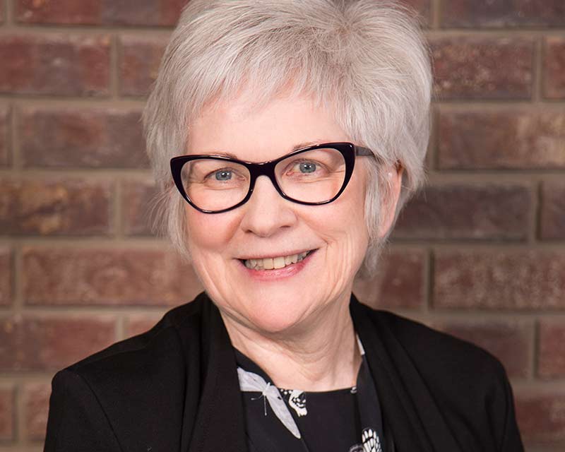 portrait of smiling woman with gray hair black rimmed glasses and black clothing against brick wall