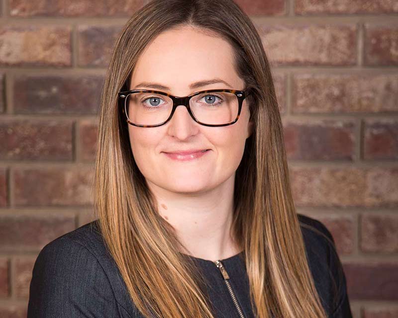 portrait of woman wearing glasses with long dirty blonde hair against brick wall